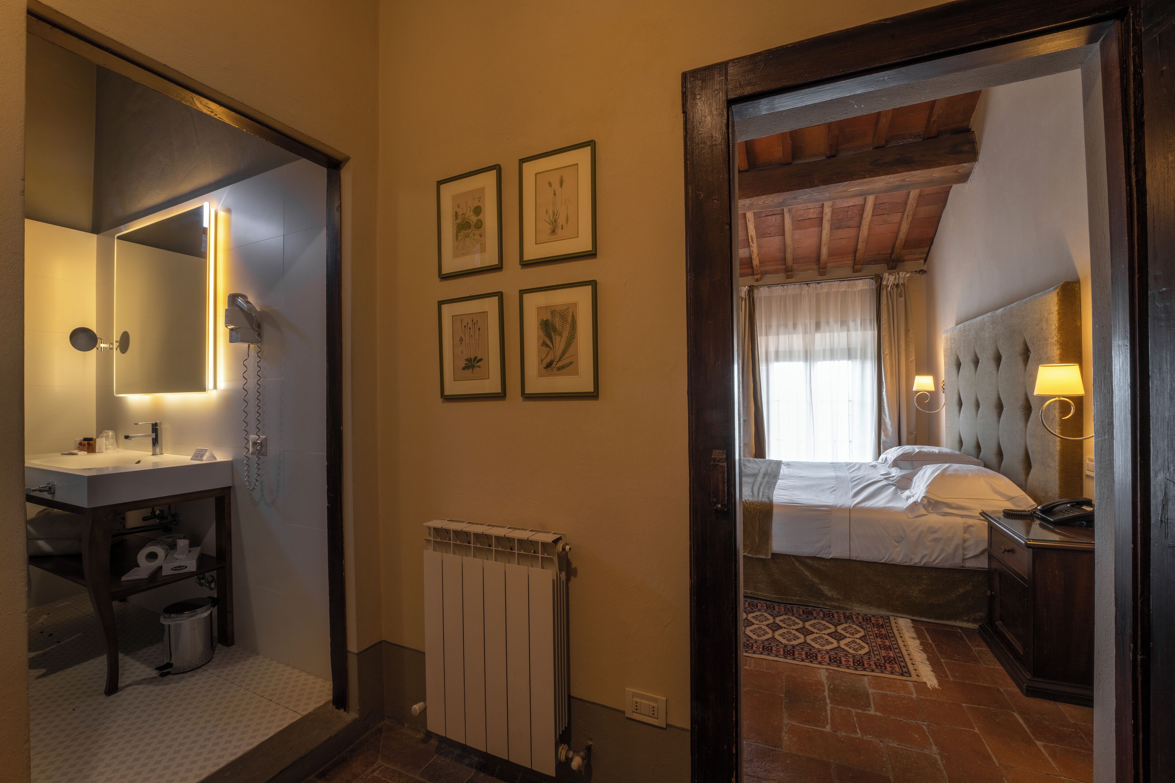 A hotel room with a bathroom, a bed with a headboard, a bedside table with a lamp, and a wall with framed pictures.