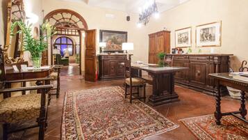 A spacious room with a large wooden desk, a chair, a table, and a cabinet. There is a mirror, a lamp, and a plant on the desk. The room has a tiled floor and a rug.