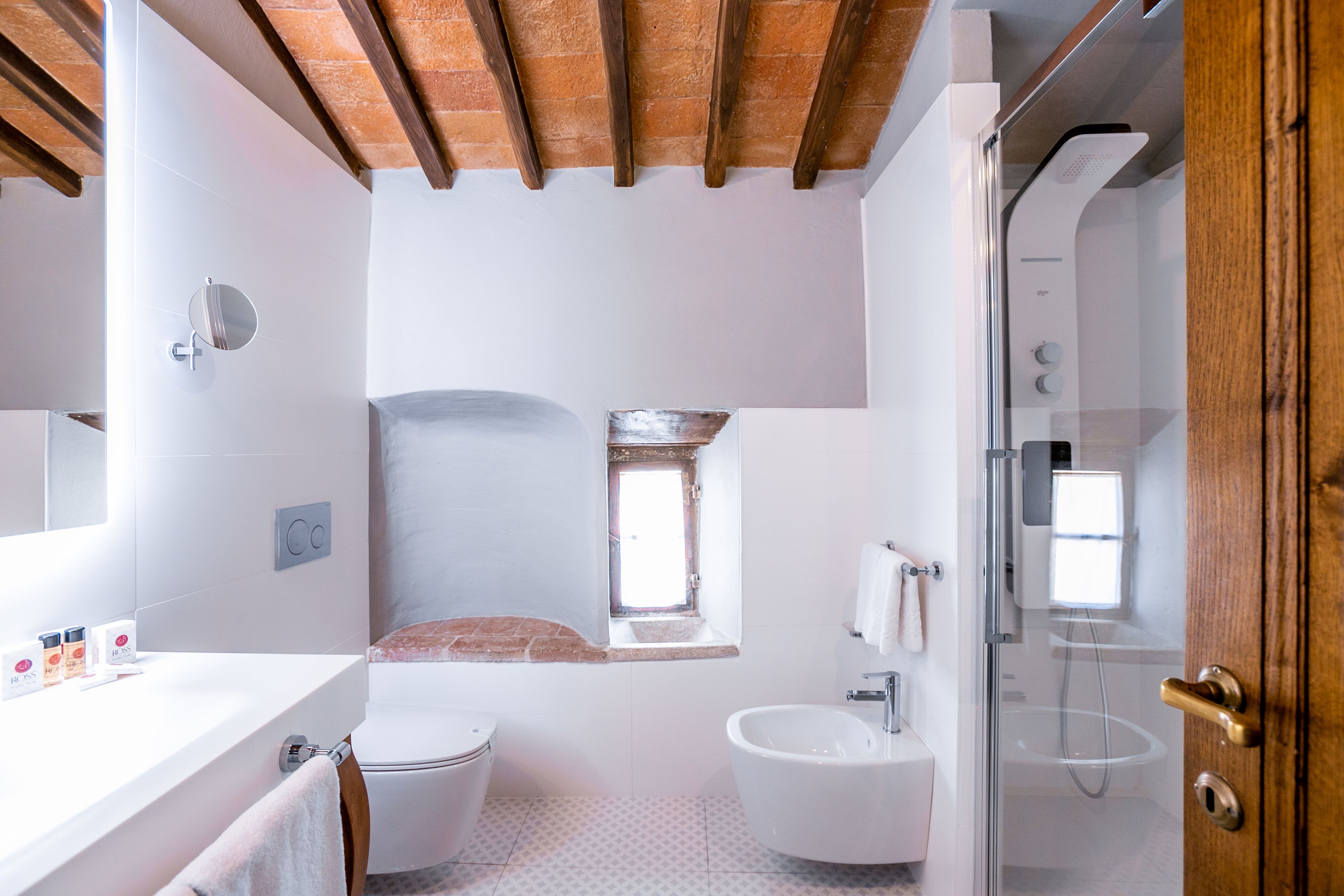 A bathroom with a toilet, sink, and shower. There is a window above the toilet, and a wooden ceiling.