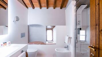 A bathroom with a toilet, sink, and shower. There is a window above the toilet, and a wooden ceiling.