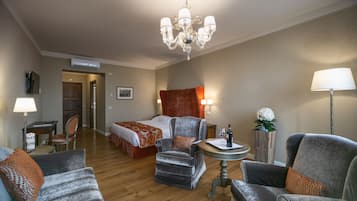 A hotel room with a bed, two armchairs, a small table, a chandelier, a lamp, and a potted plant.