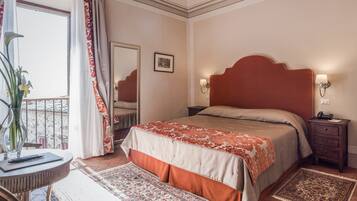 Una camera da letto con un grande letto, un lampadario, un comodino, una sedia, un tappeto, una finestra con tende e un quadro appeso al muro.