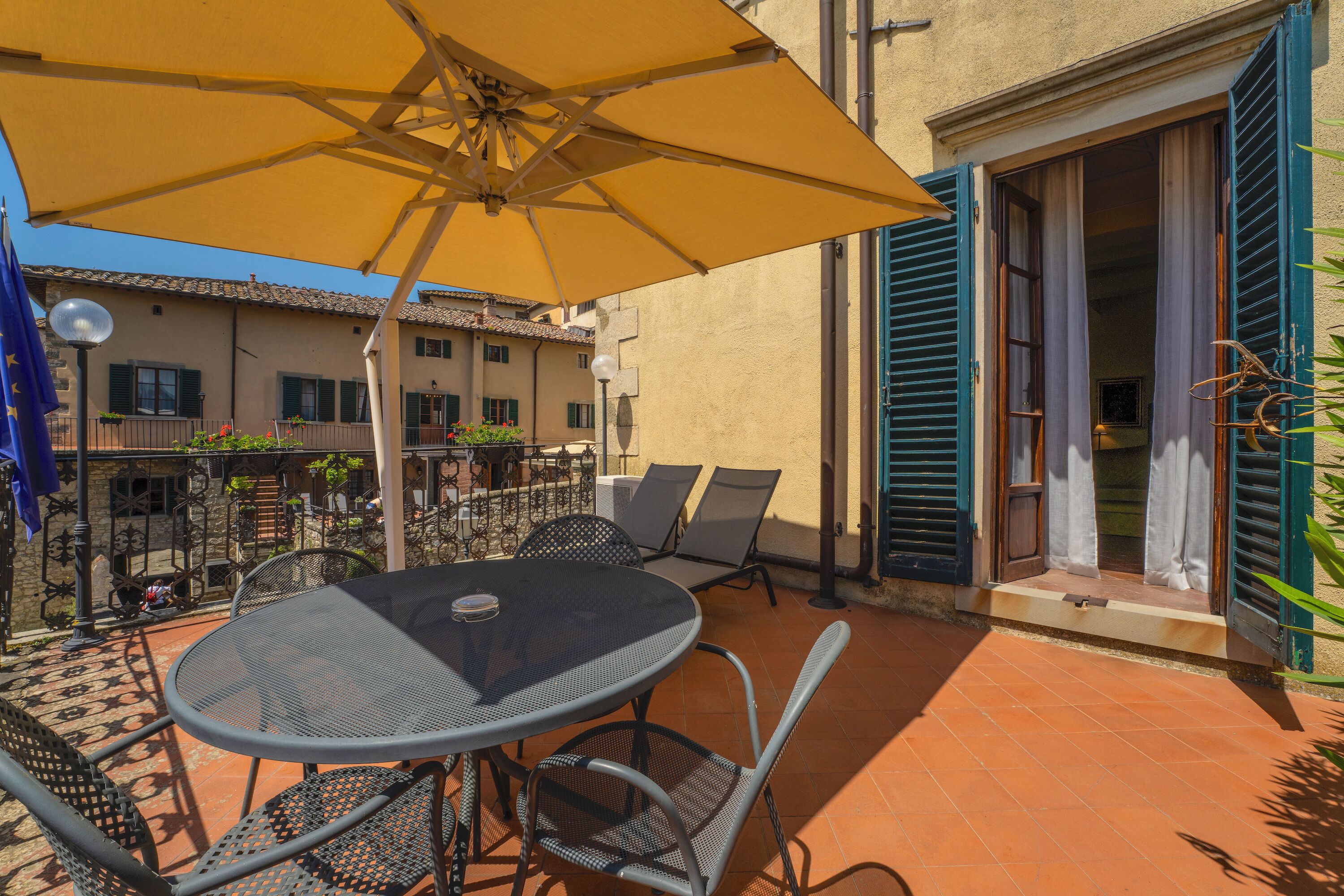 An outdoor patio with a round table and chairs, a yellow umbrella, and a building with green shutters.