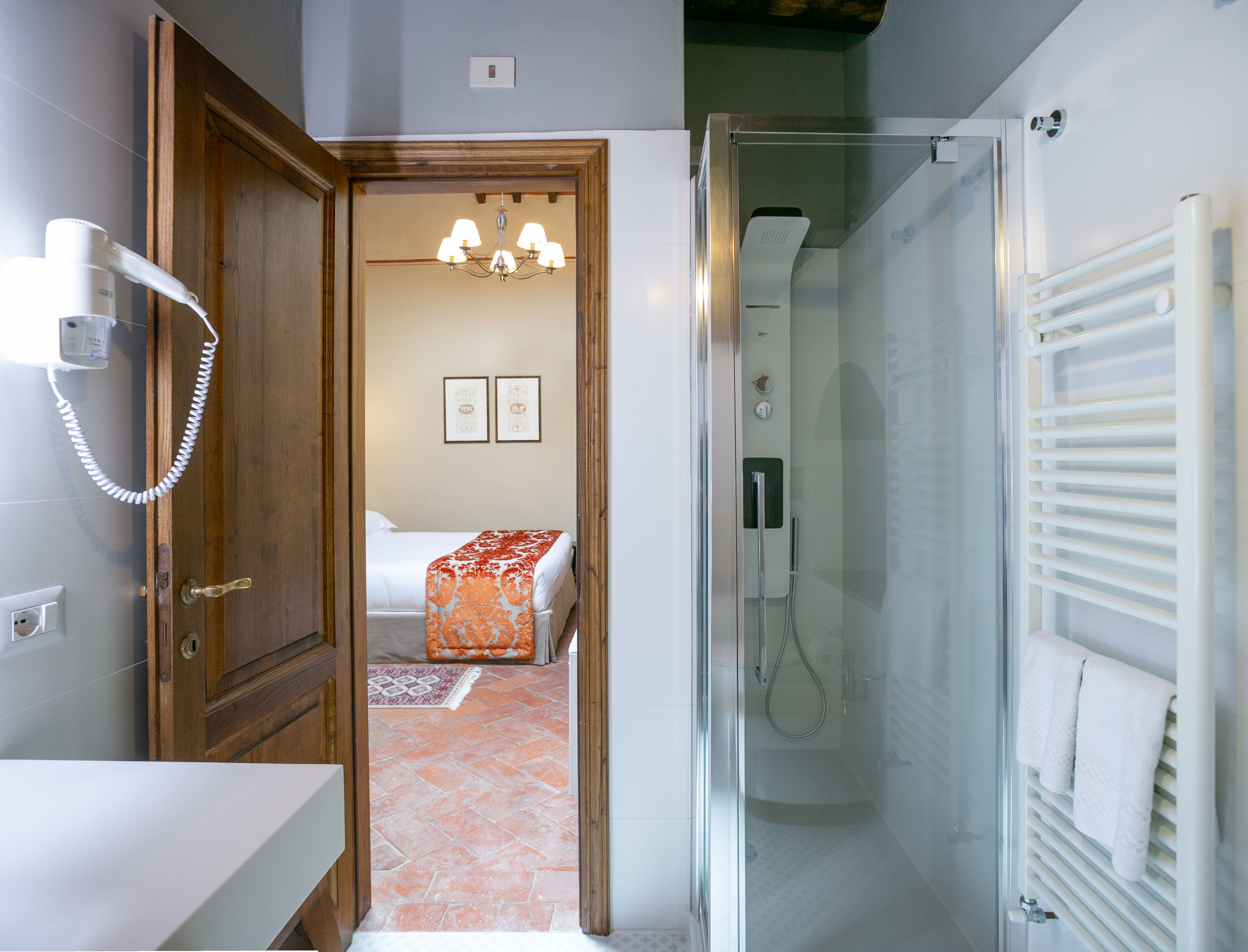 Ein Badezimmer mit Dusche, Toilette und Waschbecken. Im angrenzenden Raum befindet sich ein Bett mit Decke.