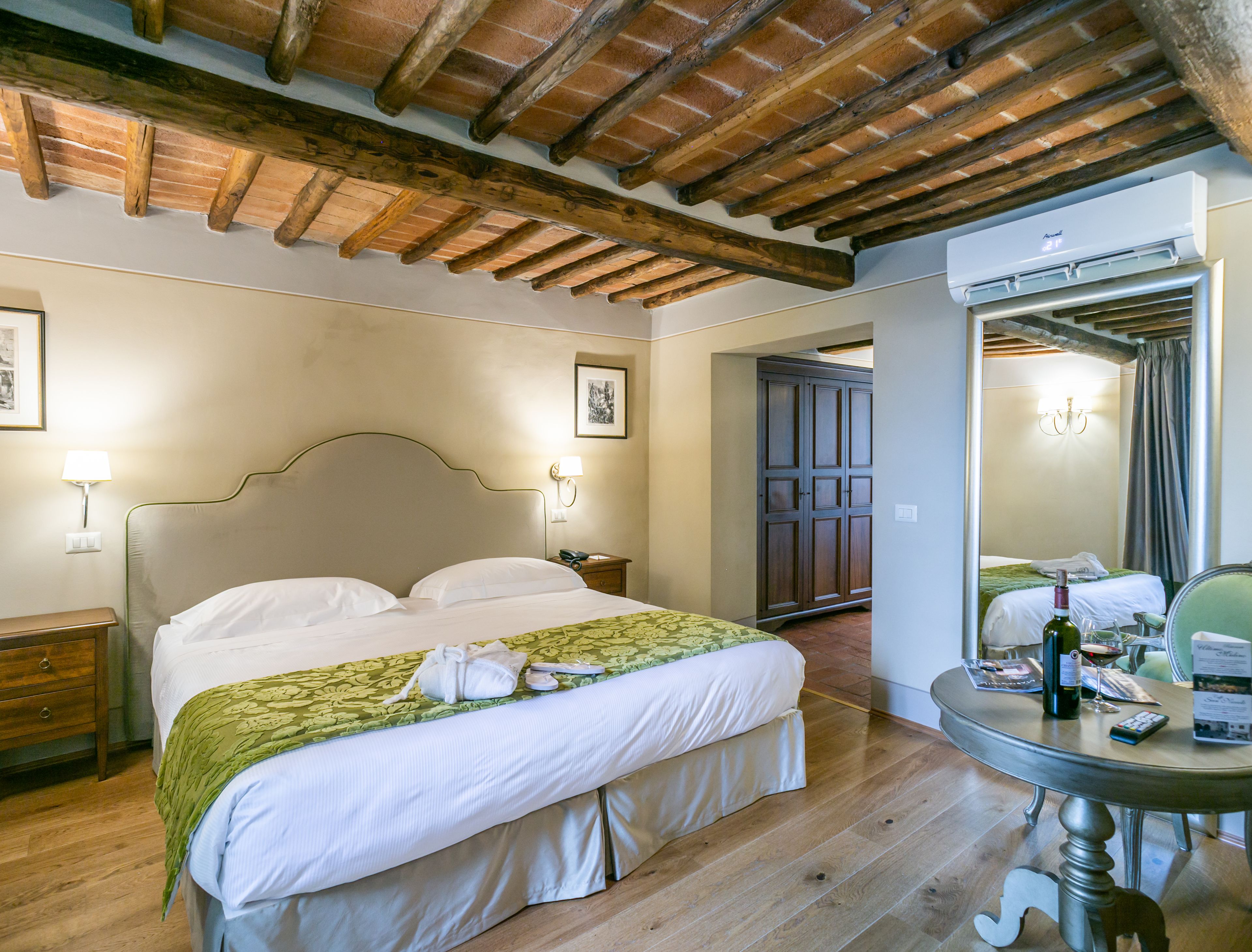 Quarto de hotel com teto de madeira, cama com colcha verde, mesa de cabeceira com abajur e um espelho grande.