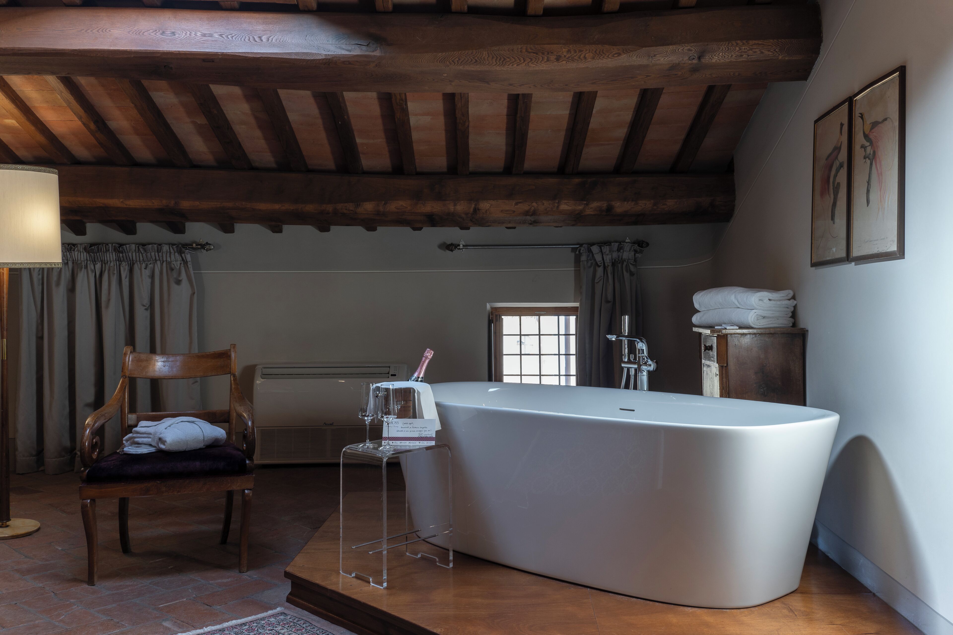 Un baño moderno con una bañera independiente, vigas de madera en el techo, una silla con toallas y una ventana.