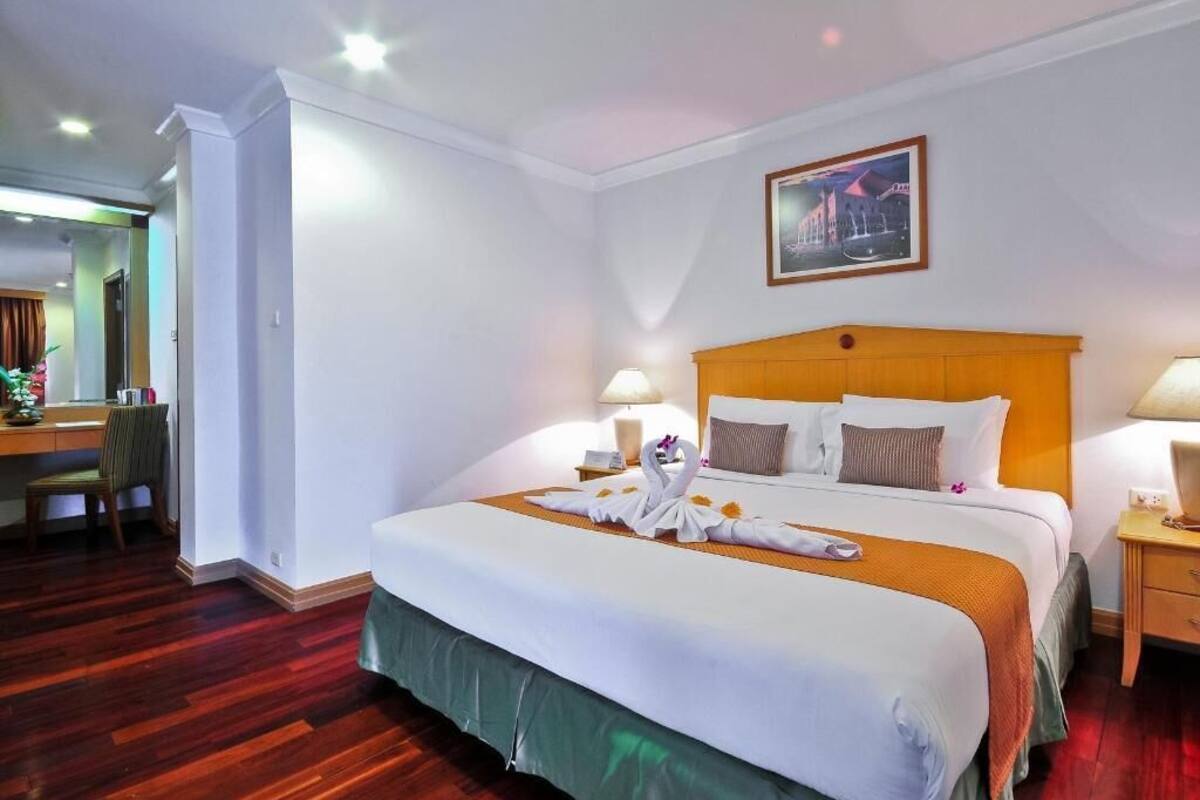 A hotel room with a large bed, a wooden headboard, two bedside tables with lamps, a desk with a chair, and a painting on the wall.