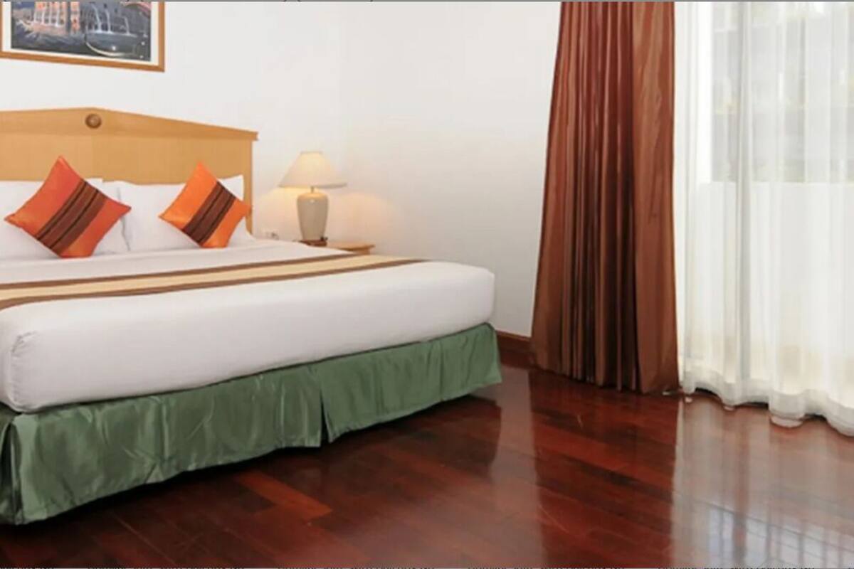 A hotel room with a wooden headboard, a bed with white bedding and green skirt, a nightstand with a lamp, and a view of a cityscape through sheer curtains.
