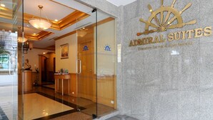 Entrada de un hotel con puerta de cristal, mostrador de recepción de madera y un cartel que dice "Admiral Suites".