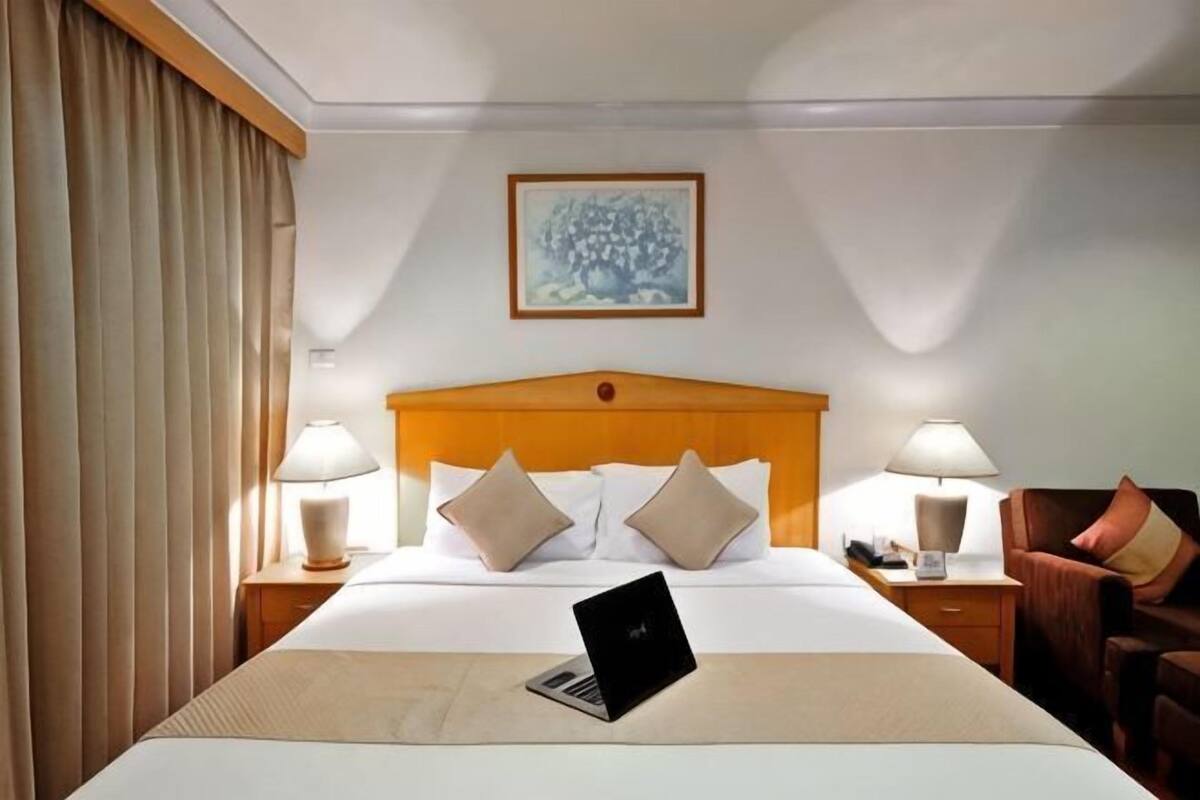A hotel room with a bed, a laptop, two bedside lamps, a wooden headboard, and a painting on the wall.
