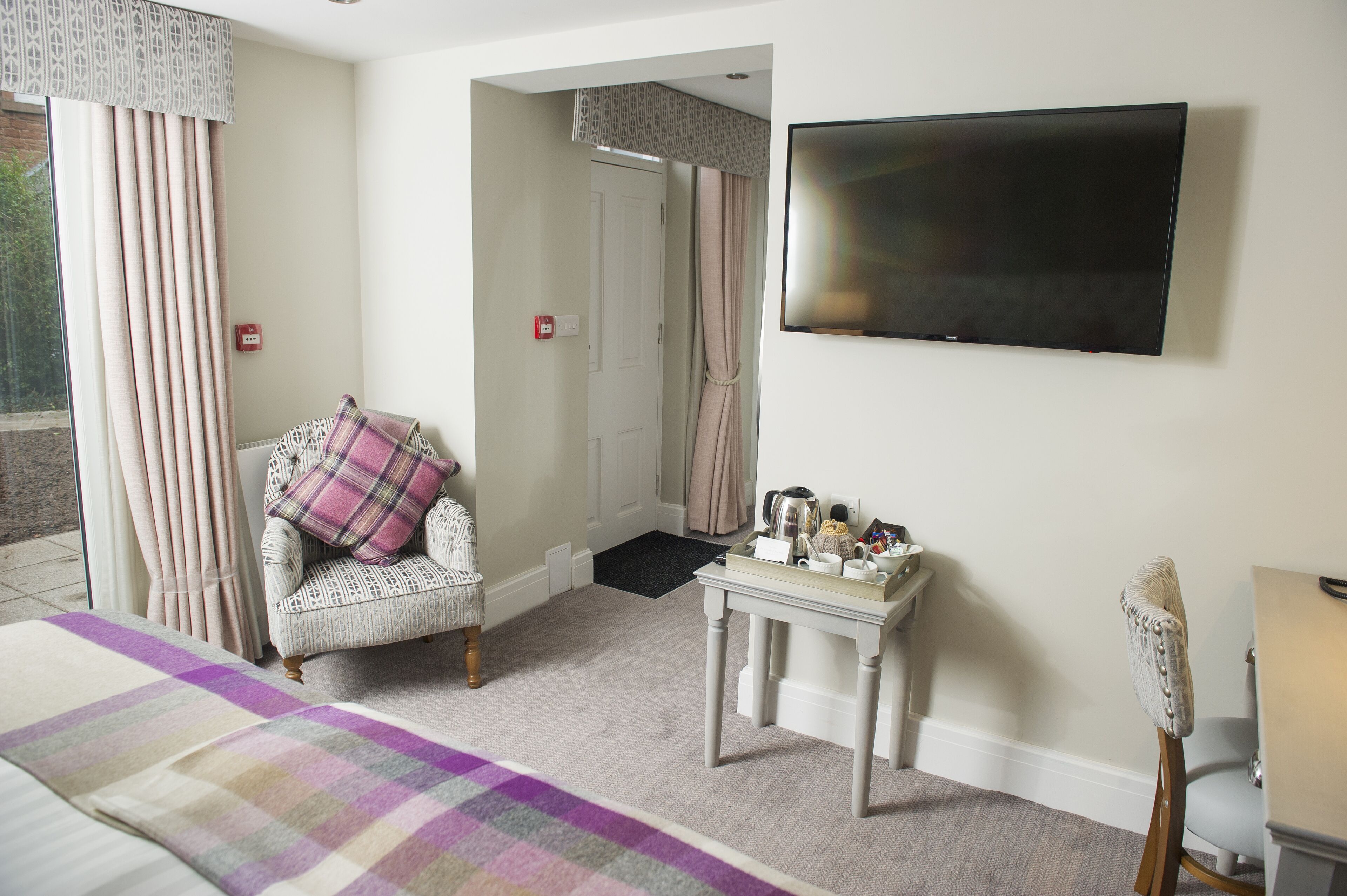 A hotel room with a bed, a chair with a plaid cushion, a small table with items, a television, and a door leading to another room.