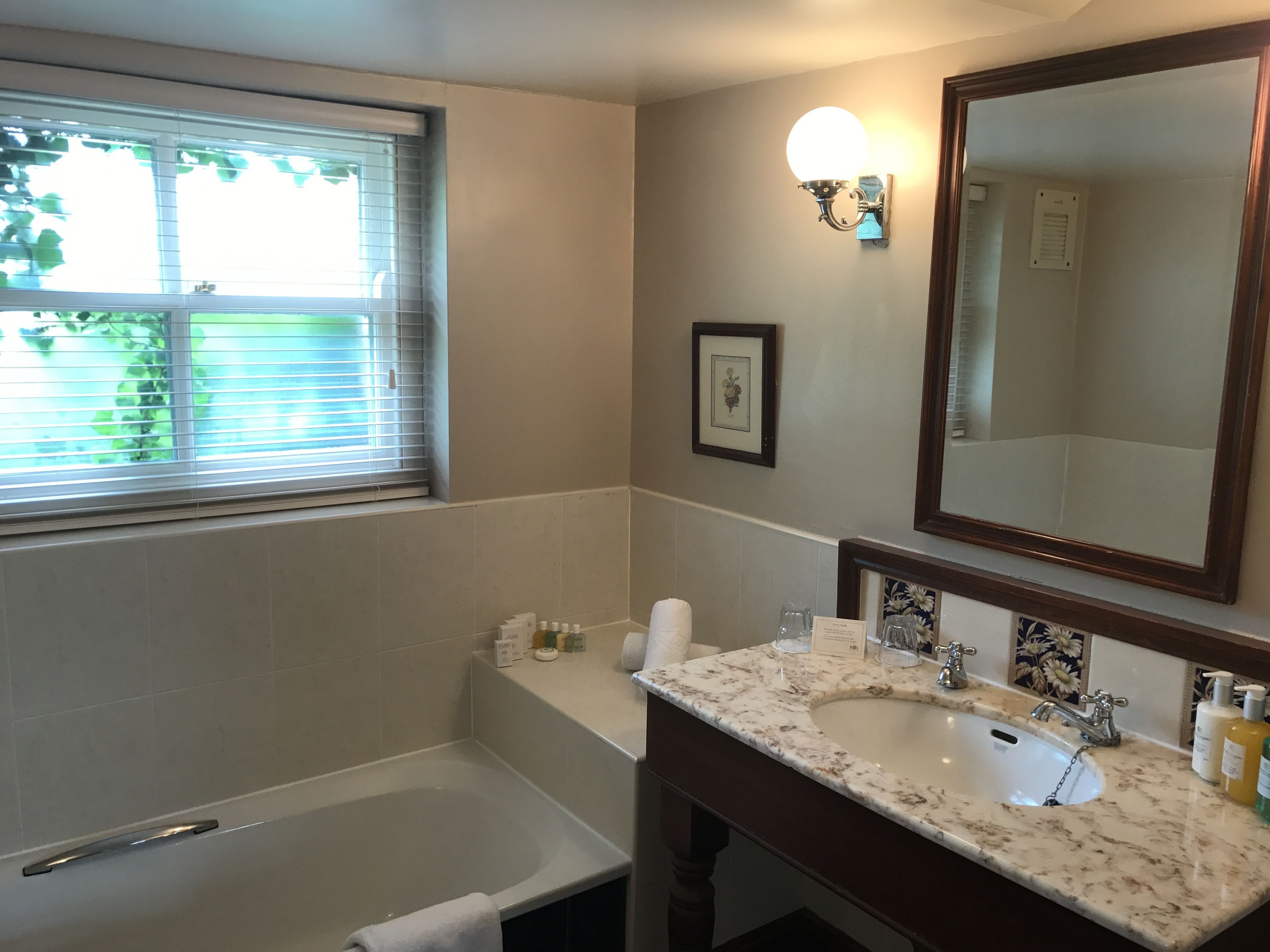 A bathroom with a bathtub, a sink with a marble countertop, a large mirror, and a window with blinds.