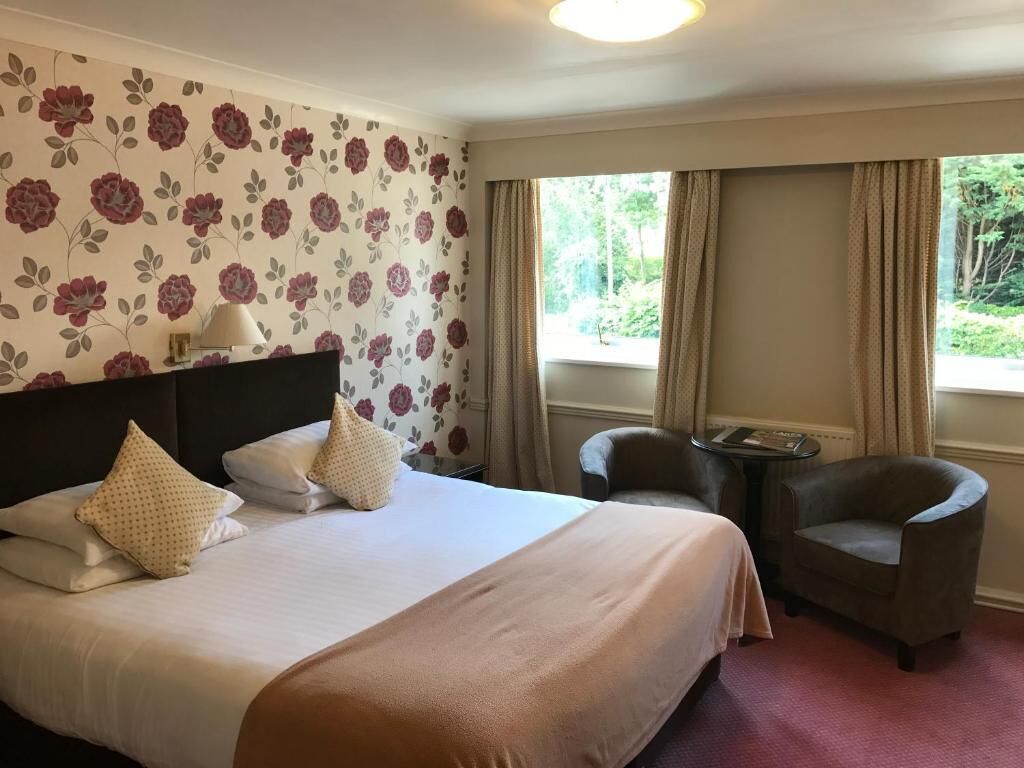 A hotel room with a large bed, two chairs, a small table, and a window with curtains.
