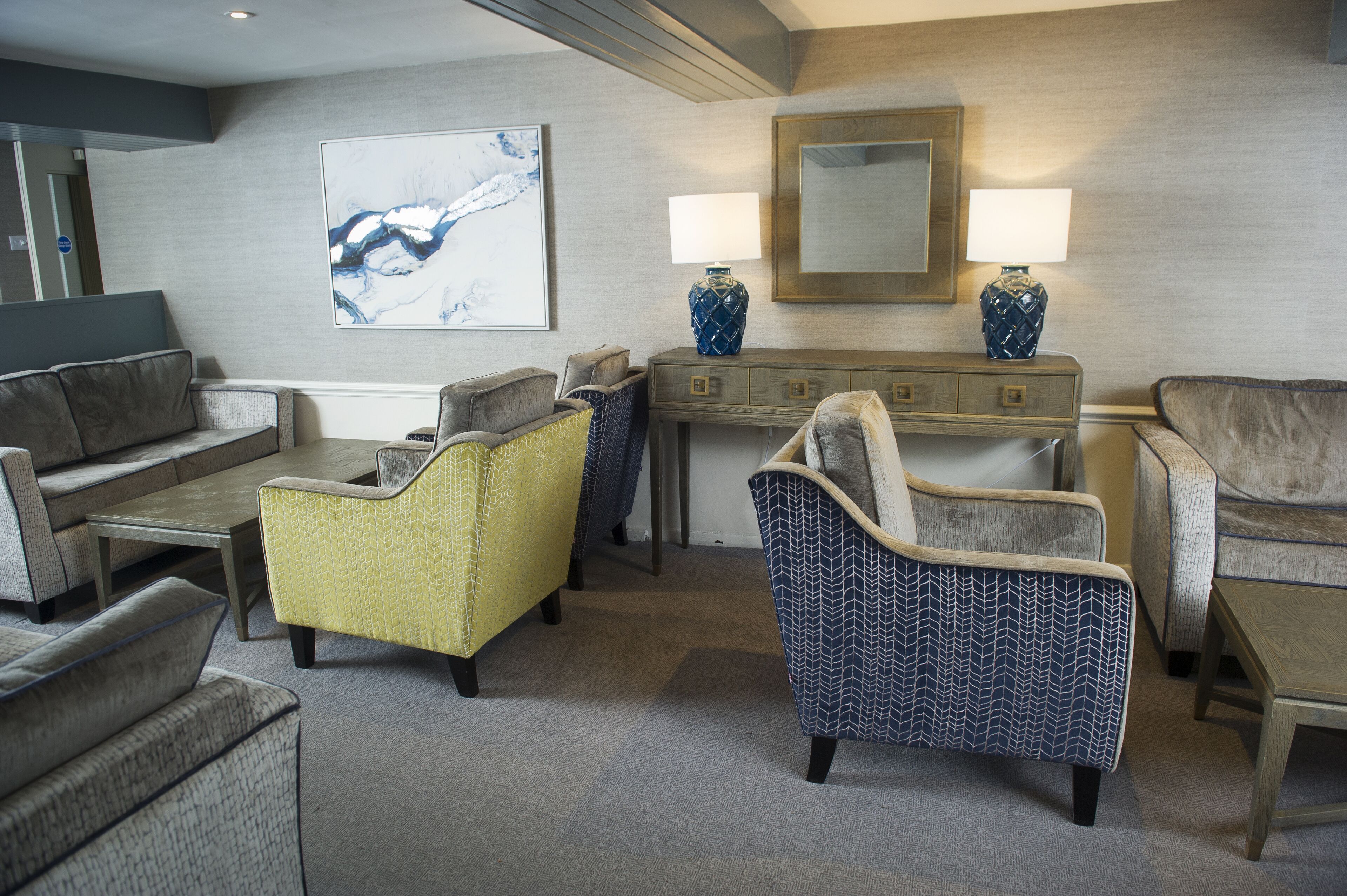 Een hotelkamer met een zithoek, fauteuils, een salontafel, een console met lampen en een groot abstract schilderij aan de muur.