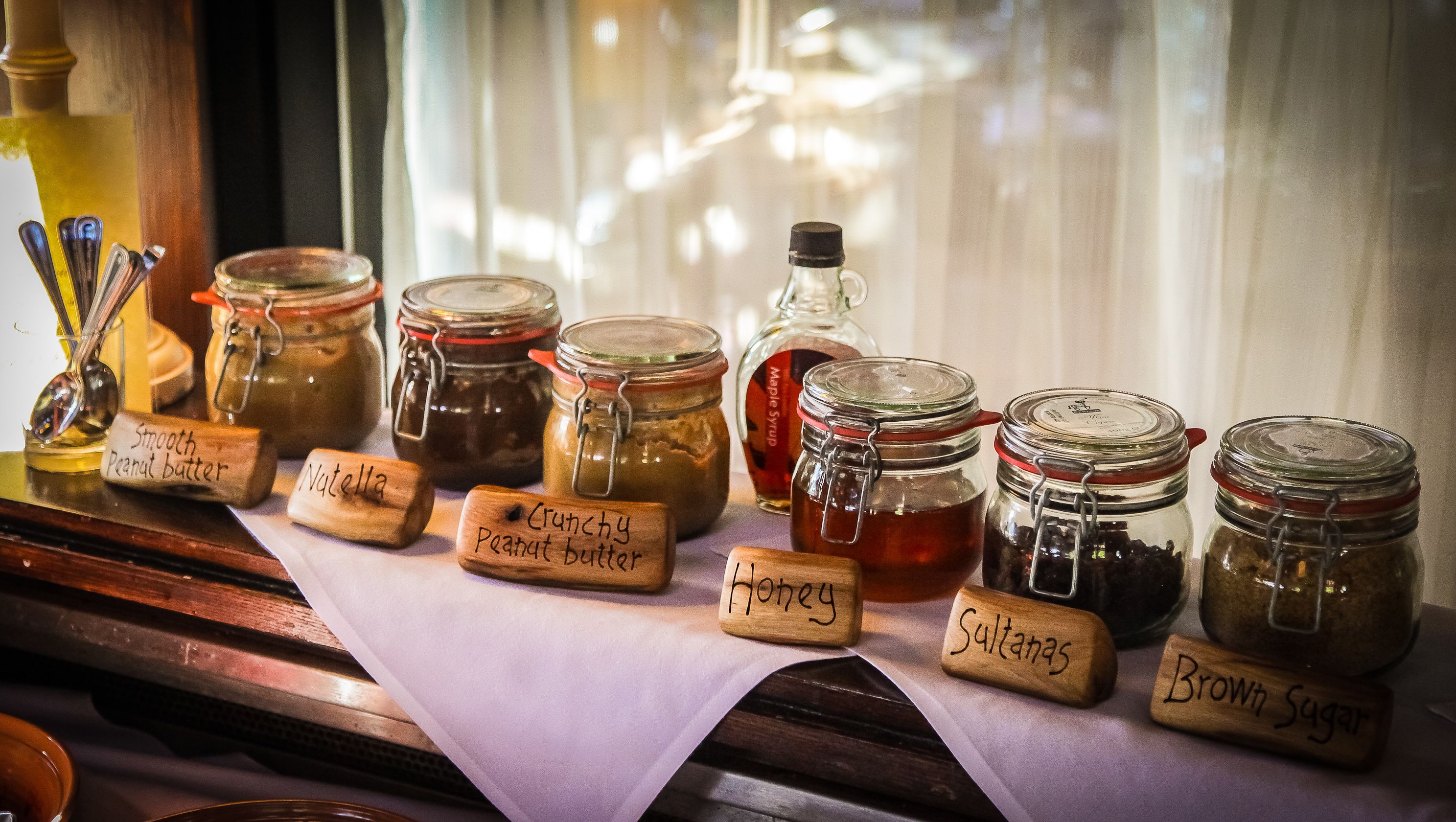 Eine Auswahl verschiedener Lebensmittel in Gläsern mit Holzschildern, darunter Erdnussbutter, Honig und brauner Zucker.