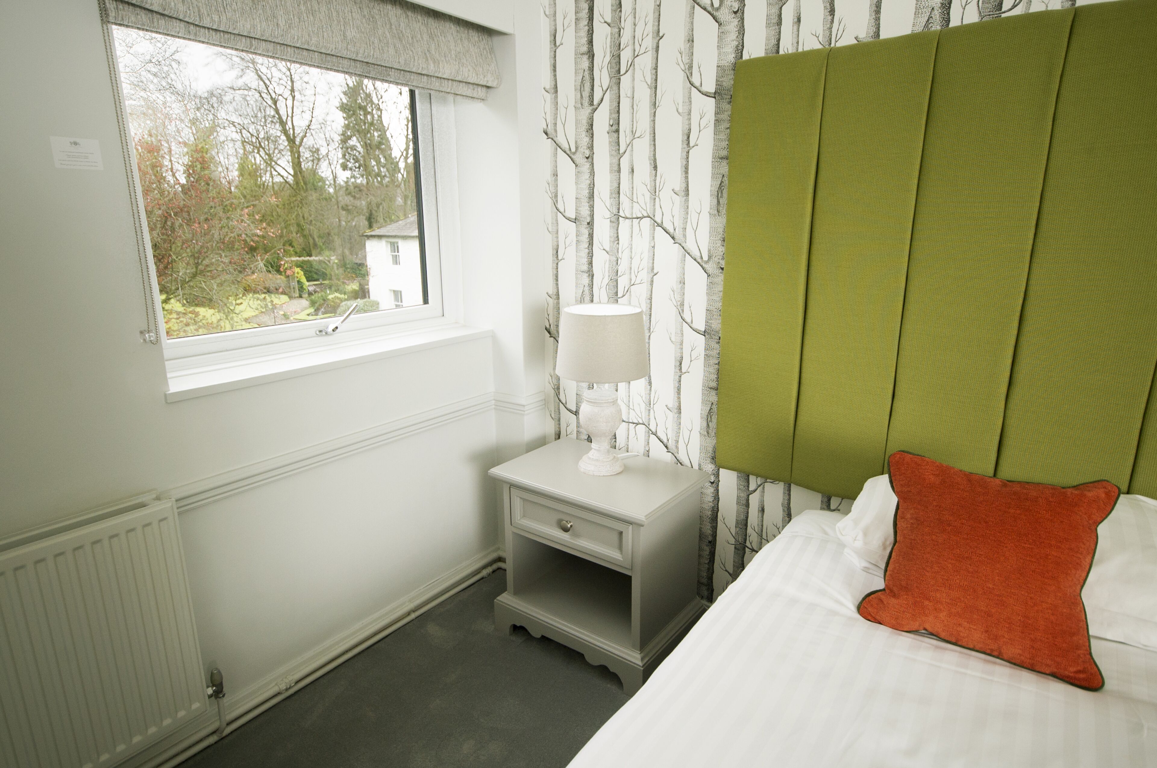 A bedroom with a window, a nightstand with a lamp, a bed with a pillow, and a headboard.