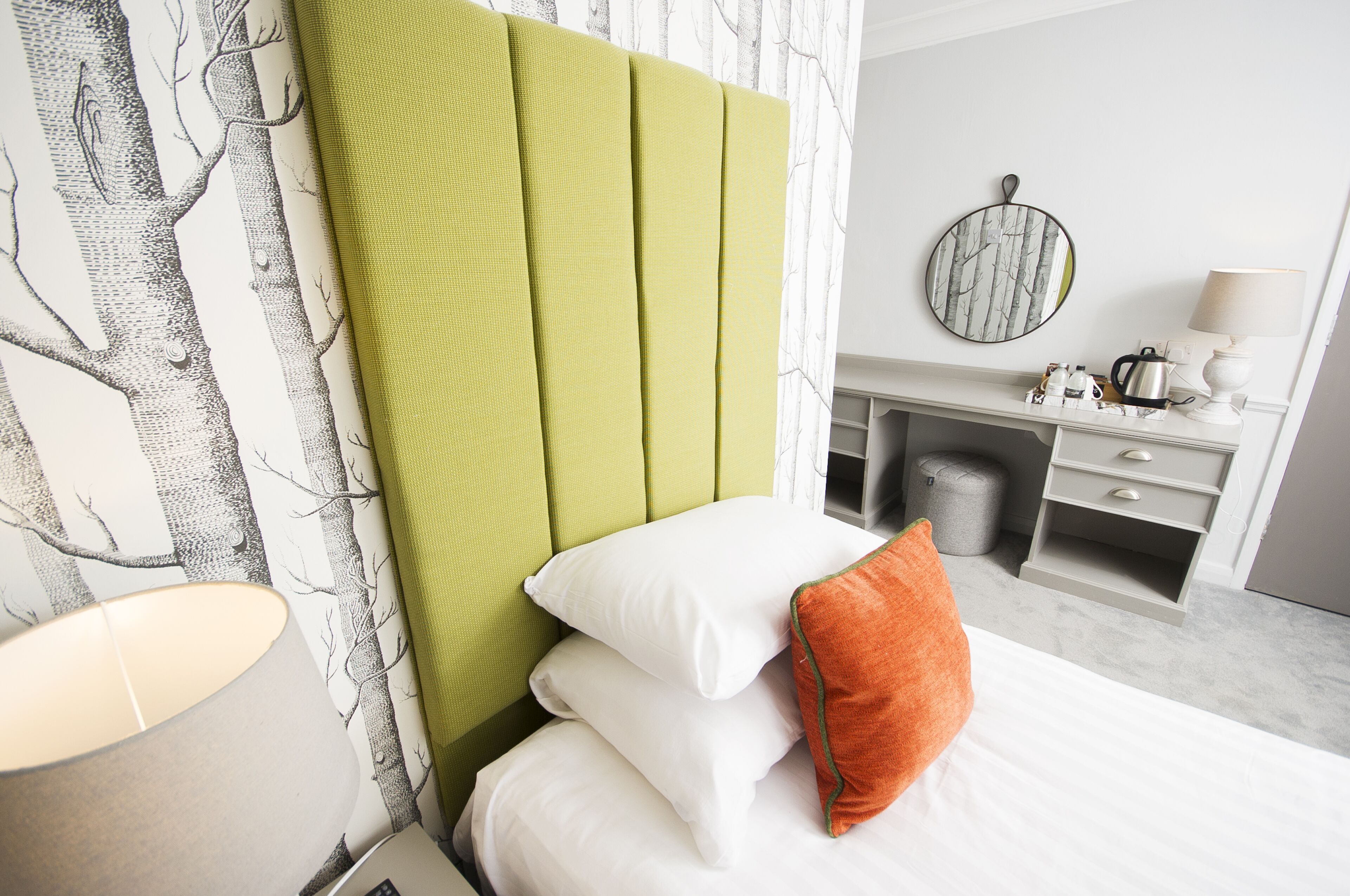 A hotel room with a green upholstered headboard, white bedding, and a round mirror on the wall.