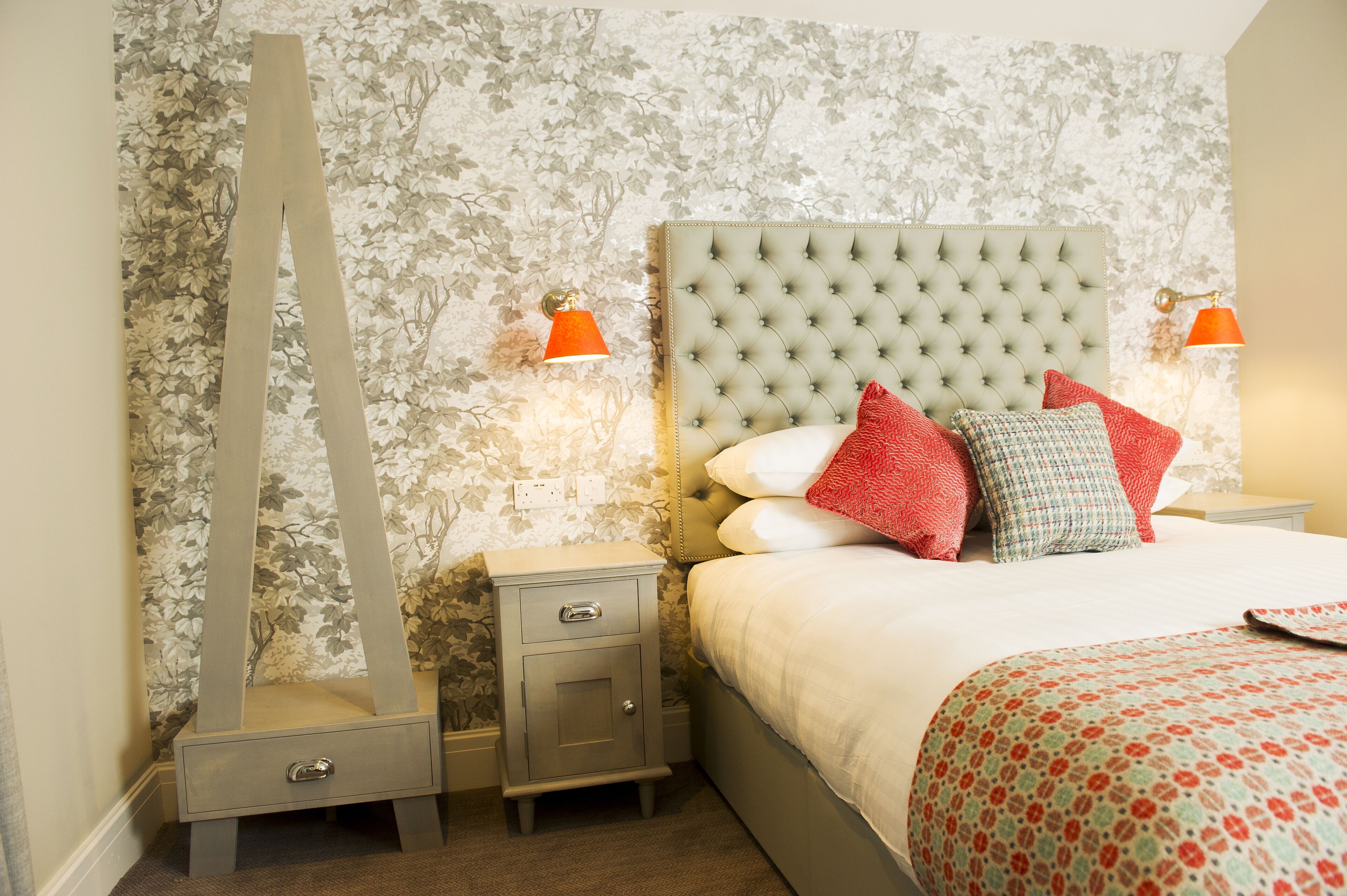 A bedroom with a tufted headboard, a bed with red and white pillows, two bedside tables with lamps, and floral wallpaper.