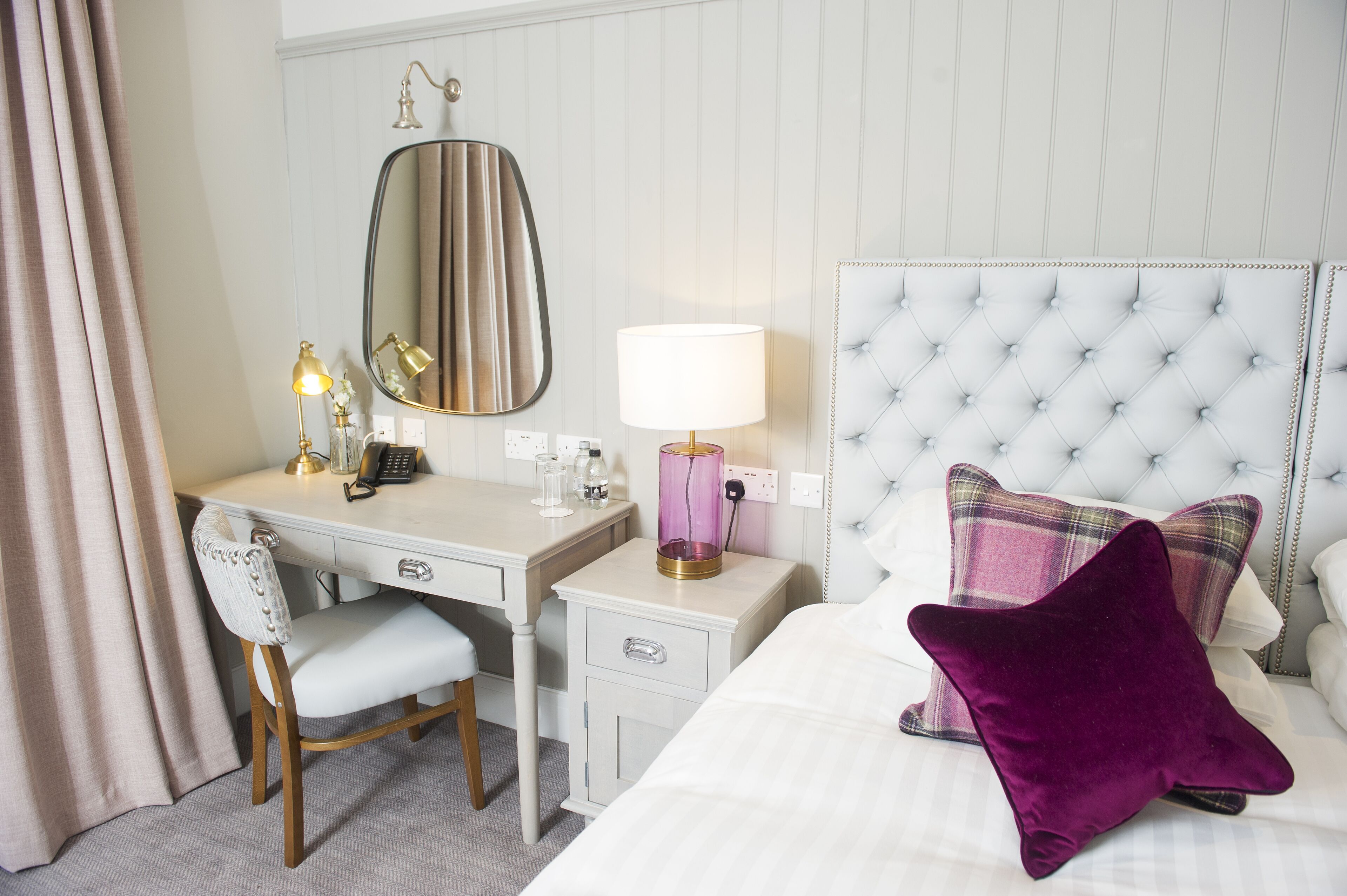 A hotel room with a bed, a desk, a chair, a vanity with a mirror, a lamp, and a bedside table.