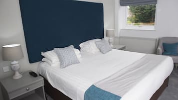 A neatly made bed with a blue headboard, white bedding, and patterned pillows. There is a bedside table with a lamp and a phone. A chair is visible in the corner, and a window with a view of greenery is in the background.