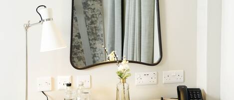 Un bureau dans une chambre d’hôtel, avec un miroir, une lampe, un vase de fleurs, un téléphone et une chaise.
