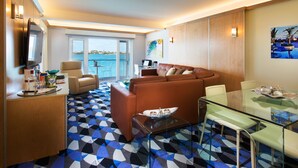Un salon moderne avec un canapé en cuir marron, une table basse en verre et une table à manger en verre, ainsi que des chaises. Une grande fenêtre offre une vue sur la mer, et il y a un balcon.