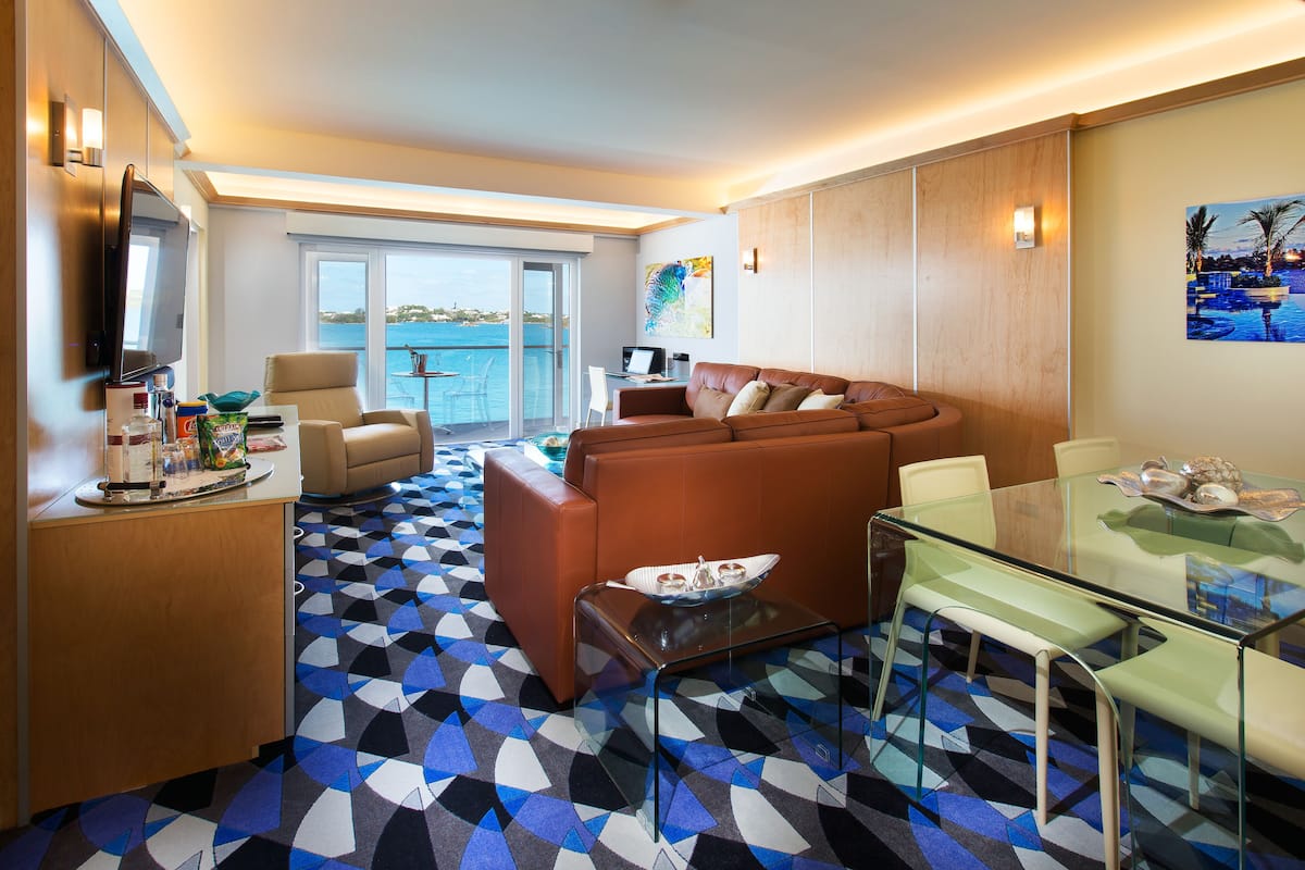 A modern living room with a brown leather sofa, a glass coffee table, and a glass dining table with chairs. There is a large window with a view of the sea and a balcony.