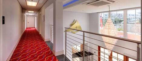 Un couloir d’hôtel avec un tapis rouge, un escalier doté d’une rampe en métal et un lustre.