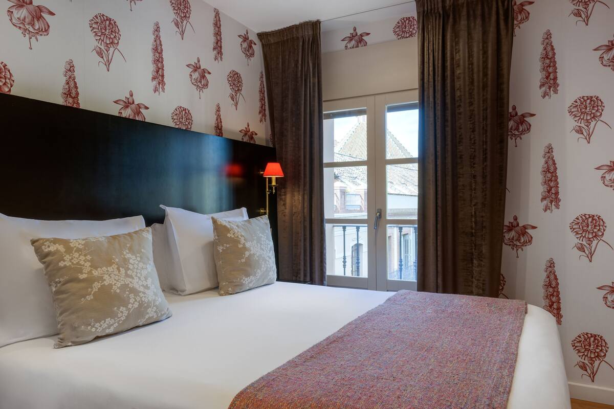 A hotel room with a large bed, floral wallpaper, and a view of a building through the window.