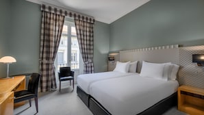 Ein Hotelzimmer mit einem großen Bett, einem Schreibtisch mit Stuhl, einem Fenster mit Vorhängen und einem Nachttisch mit Lampe.