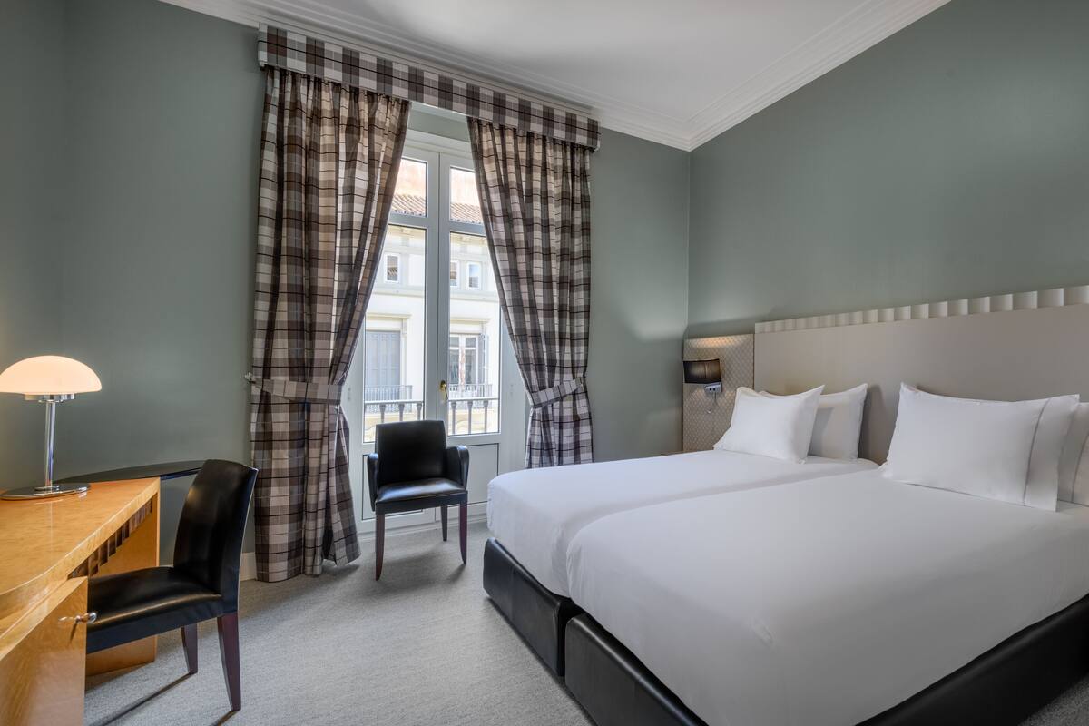 A hotel room with a large bed, a desk with a chair, a window with curtains, and a nightstand with a lamp.