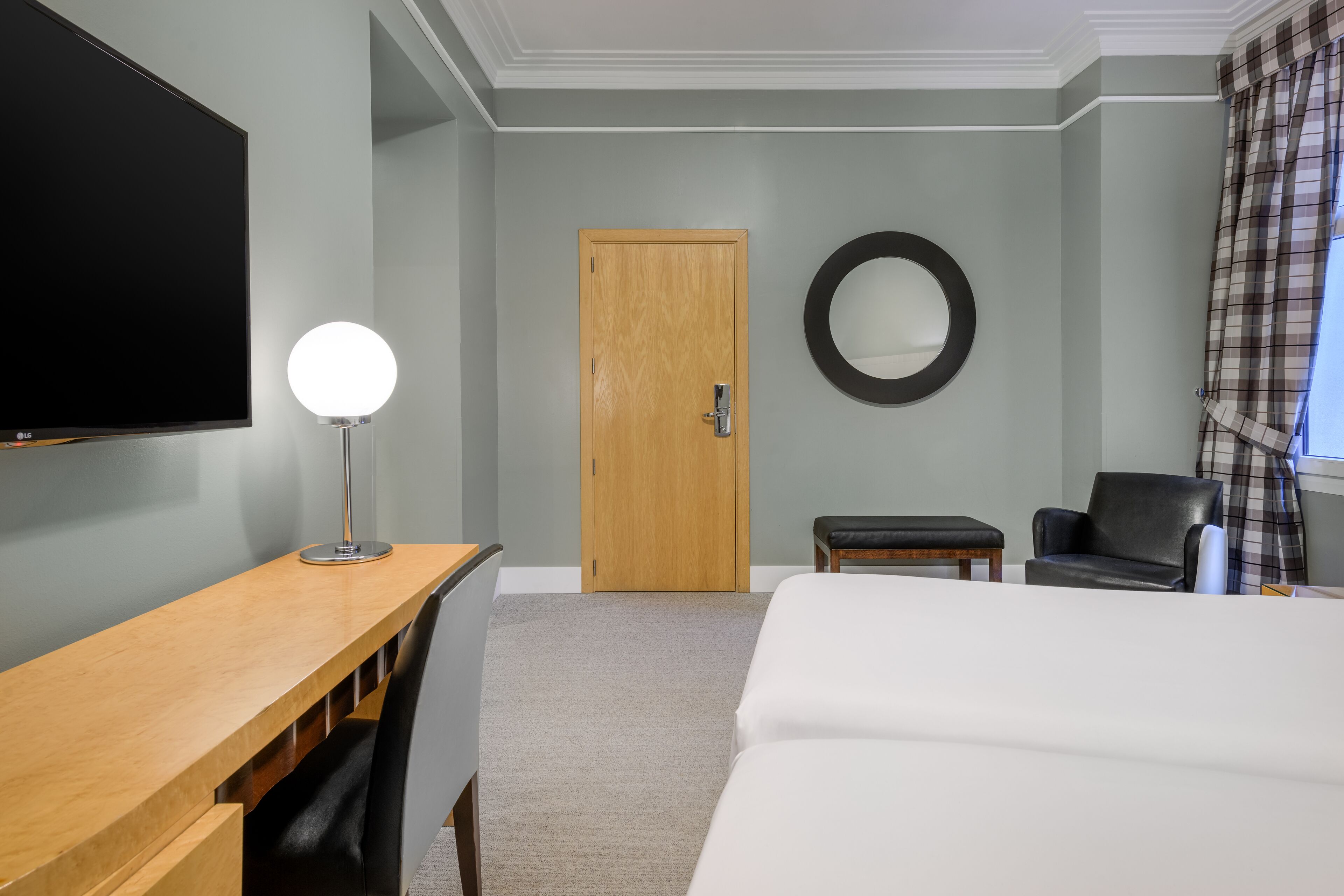 A hotel room with a bed, a desk with a lamp, a chair, a round mirror, and a door.