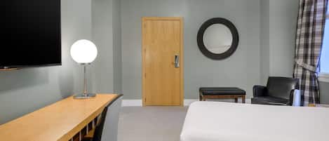 A hotel room with a bed, a desk with a lamp, a chair, a round mirror, and a door.