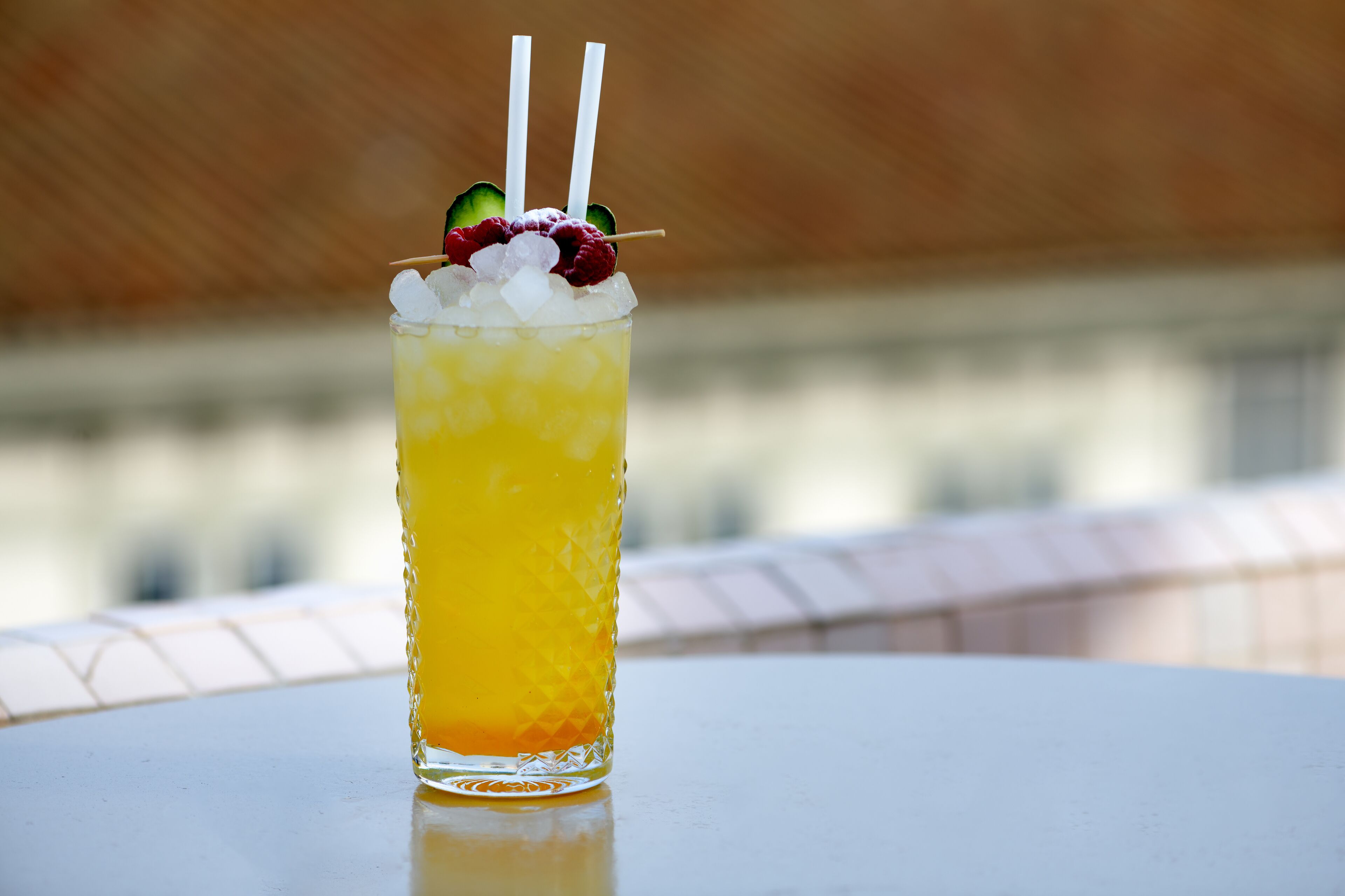 A tall glass with a yellow beverage, ice cubes, and garnishes like a slice of avocado, raspberries, and a cherry, with two straws.