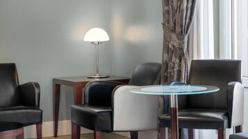 A hotel room with a seating area featuring two black leather chairs, a white chair, and a small round table with a glass top. There is a floor lamp with a white shade and a patterned curtain on the window.