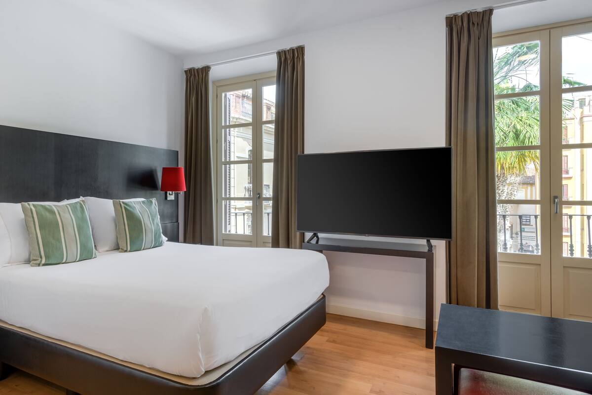 A hotel room with a large bed, a TV, and a desk. There are windows with curtains, and a view of buildings outside.