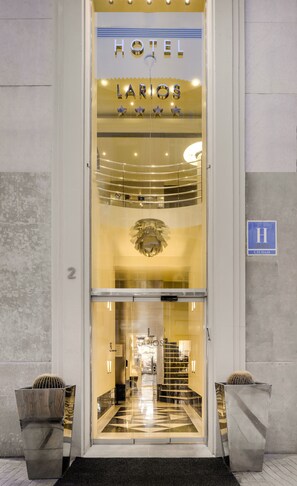Entrada de un hotel con diseño moderno, que presenta una puerta de cristal, una araña y un letrero que dice "HOTEL LARIOS".