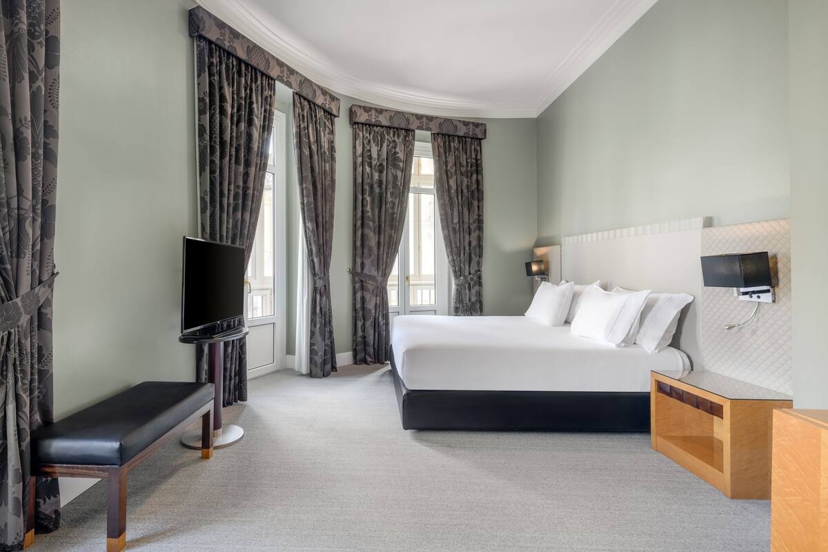 A hotel room with a large bed, a TV, a bench, and a desk.
