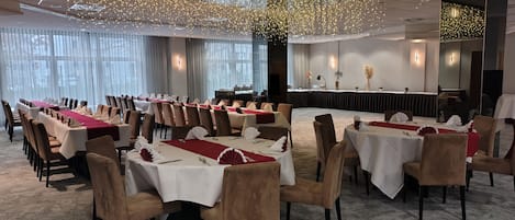 Un salón de conferencias con mesas dispuestas para una reunión, un escenario con atril y un amplio ventanal con cortinas.