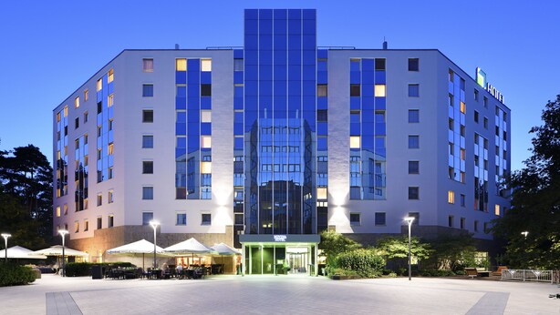 Un hotel moderno con fachada de vidrio central, varios pisos y una entrada bien iluminada.