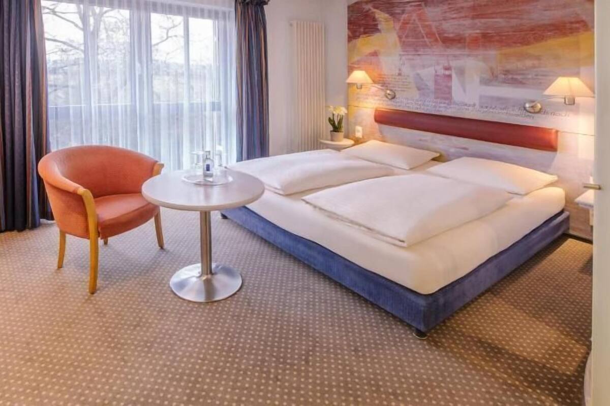 A hotel room with a bed, a chair, a small table, and a window with curtains.