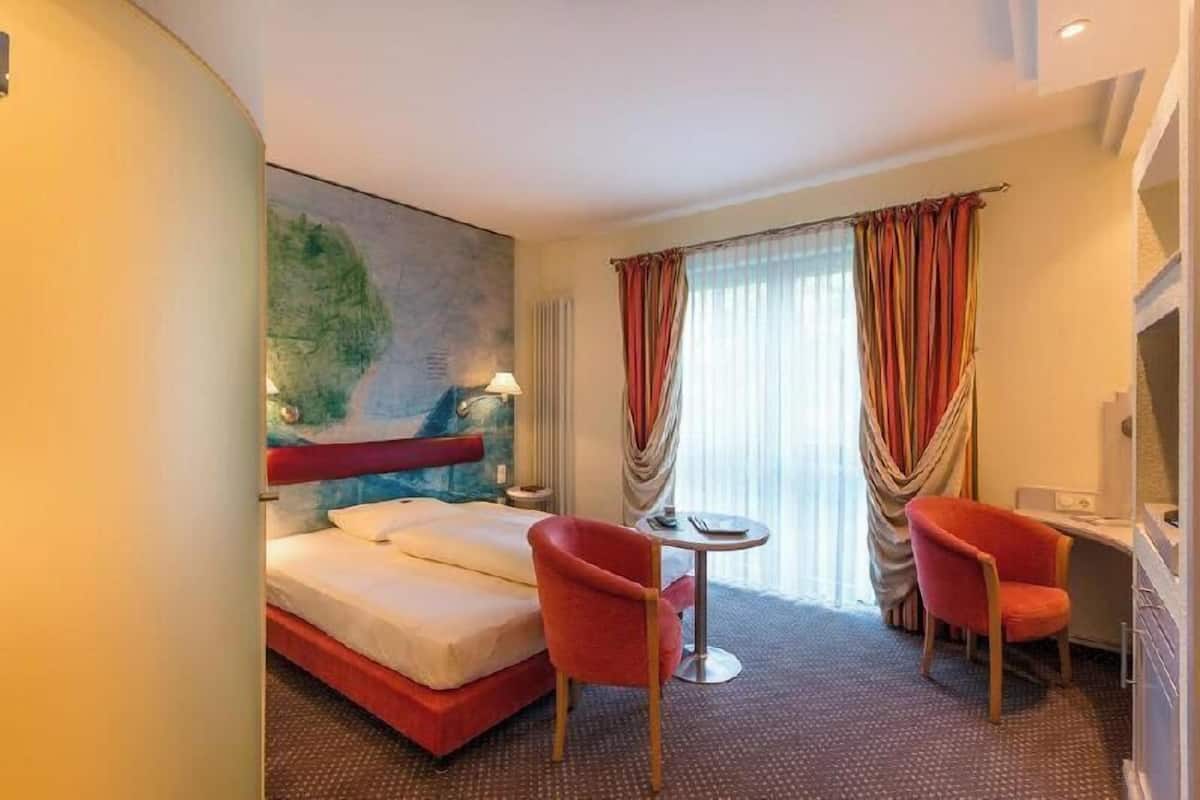 A hotel room with a bed, two chairs, a small table, a painting on the wall, and a window with curtains.