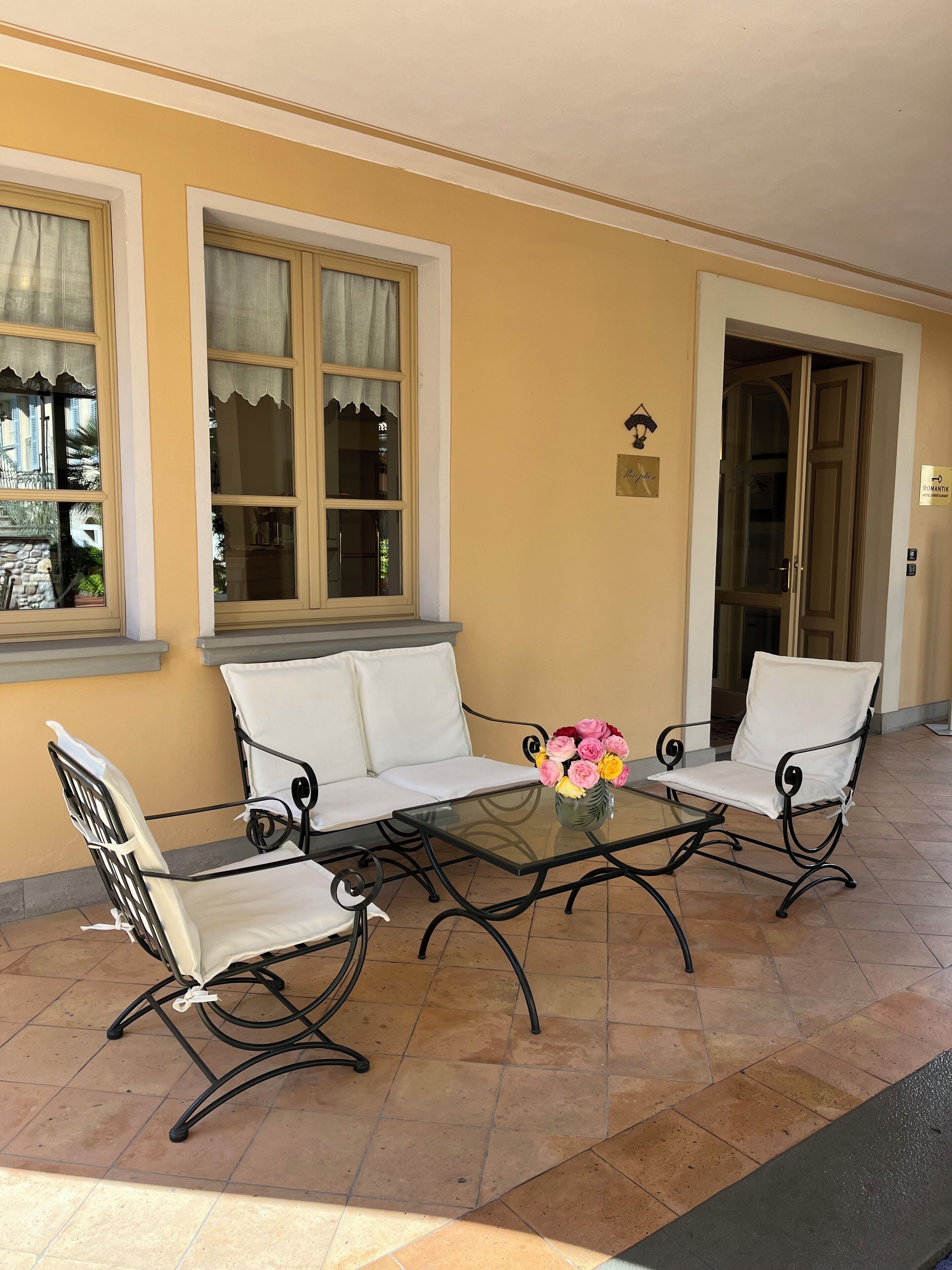 Un patio con una mesa de vidrio, sillas blancas acolchadas y un jarrón de flores.