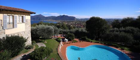 Una piscina con vistas a montañas y un lago, rodeada de vegetación y un edificio de piedra.