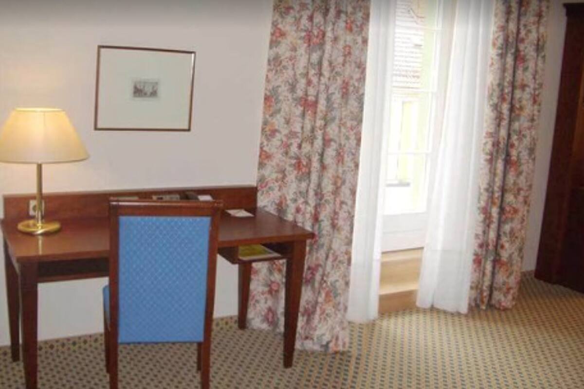 Una habitación de hotel con un escritorio de madera, una silla azul, una lámpara, un cuadro en la pared y cortinas con estampado floral.