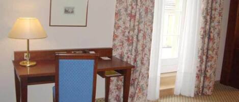 Ein Hotelzimmer mit einem Holztisch, einem blauen Stuhl, einer Lampe, einem gerahmten Bild an der Wand und blumigen Vorhängen.
