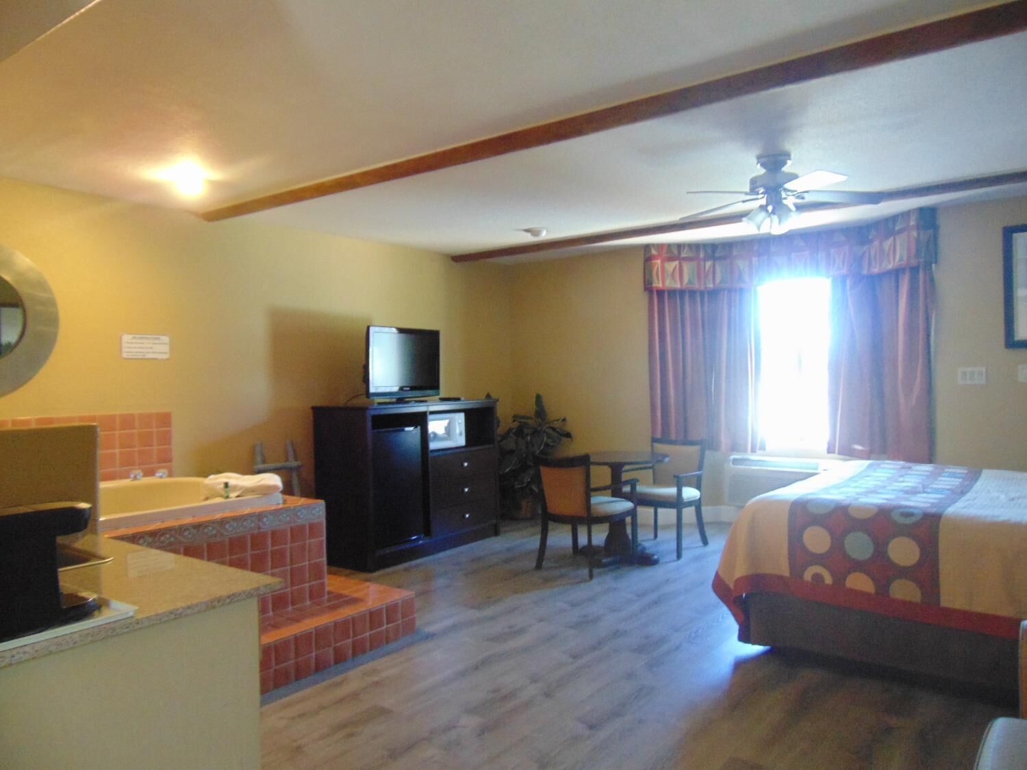 A hotel room with a bed, a desk with a computer, a television, a bathroom with a bathtub, and a ceiling fan.