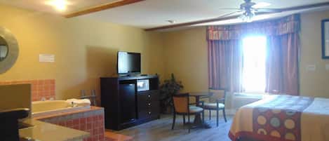 A hotel room with a bed, a desk with a computer, a television, a bathroom with a bathtub, and a ceiling fan.