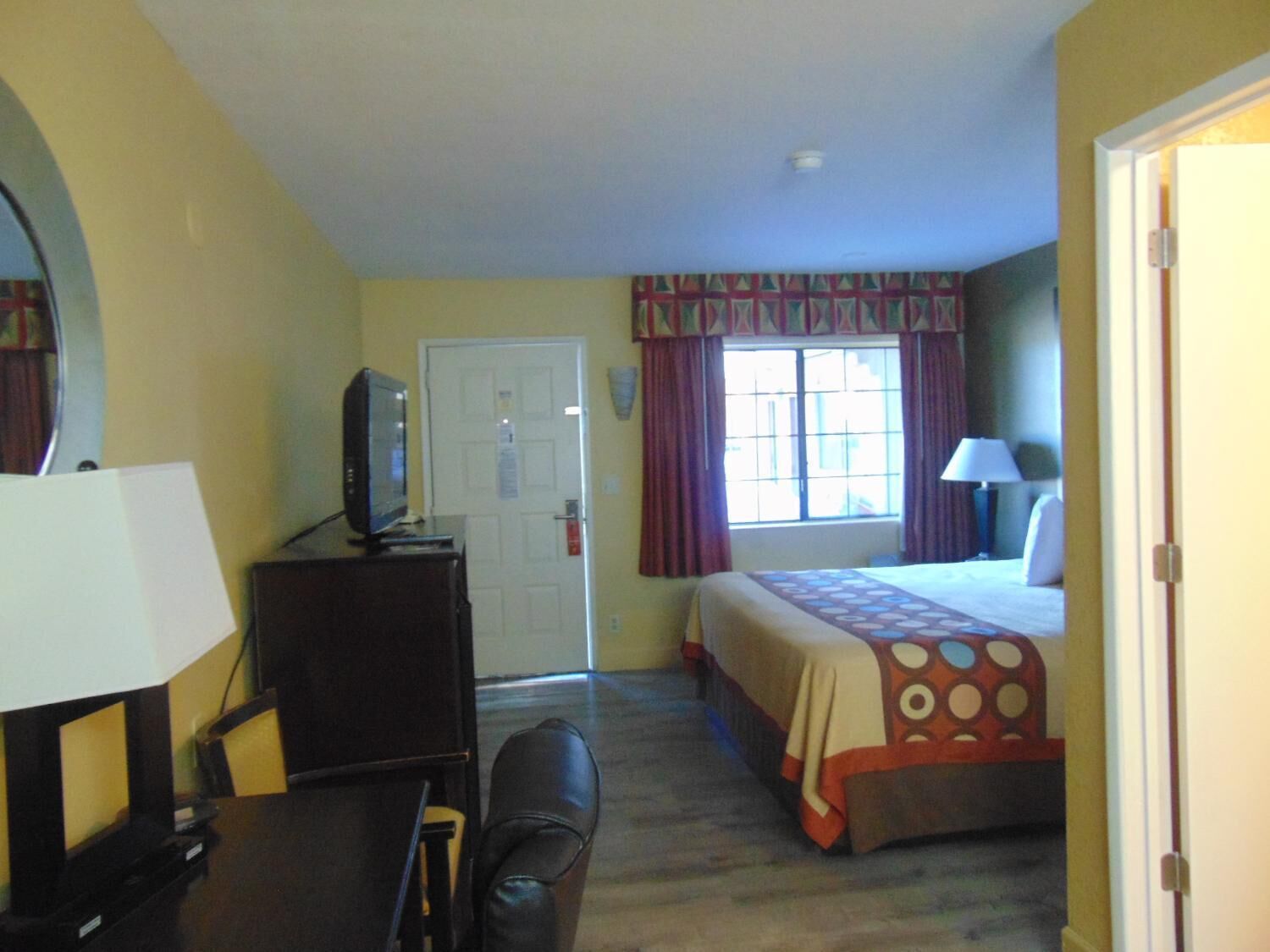 A hotel room with a bed, a desk with a television, a chair, a window with curtains, and a door.