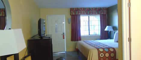 A hotel room with a bed, a desk with a television, a chair, a window with curtains, and a door.